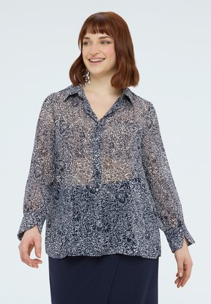 Uśmiechnięta kobieta o rudych włosach w przezroczystej, długorękawowej bluzce w kwiaty i granatowej spódnicy na jednolitym tle.