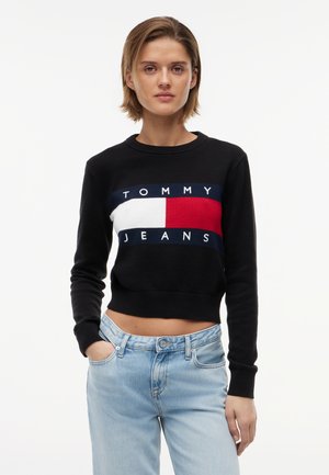 Kvinne iført en svart kort genser med en rødt, hvitt og marineblått Tommy Jeans-logo, kombinert med lyseblå denimjeans, stående med hendene i lommene.