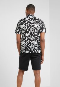 Man i en svart och vit abstrakt mönstrad kortärmad skjorta och svarta shorts, som står vänd bort från en helt vit vägg.