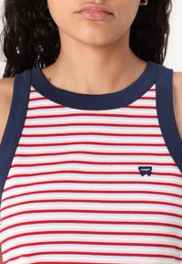Juostuotas tanko viršus su raudonomis ir baltomis horizontaliomis juostelėmis, papuoštas tamsiai mėlynomis akcentais aplink kaklo ir rankovių išpjovas. Parodytas Wrangler logotipo pleistras.