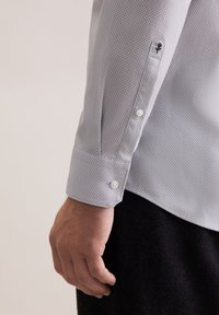 Camicia grigia chiaro con un piccolo motivo geometrico, polsini a bottone e bottoni bianchi. Presenta un accento ricamato nero sulla manica.