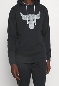 Felpa nera con logo di testa di toro bianco sul petto, cordini bianchi, indossata da una persona con pelle scura, abbinata a pantaloni neri.