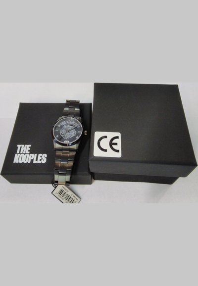 Reloj de acero inoxidable con una esfera oscura que presenta un diseño gráfico, sujetado a una pulsera metálica. Viene en dos cajas negras con etiquetas.