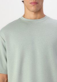 Jeune personne portant un T-shirt gris clair à col rond et manches courtes sur un fond blanc uni.