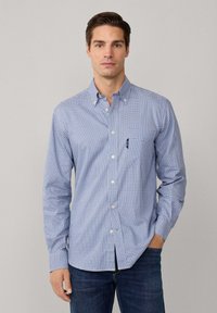 Camisa de cuadros azul y blanca de manga larga con cuello abotonado, que presenta un bolsillo en el pecho y un corte slim. Llevada con jeans oscuros.