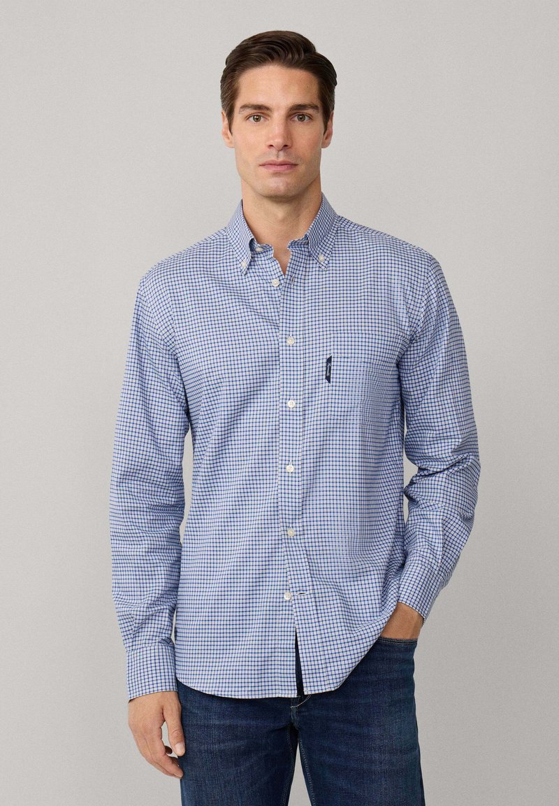 Camisa de cuadros azul y blanca de manga larga con cuello abotonado, que presenta un bolsillo en el pecho y un corte slim. Llevada con jeans oscuros.