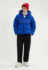 Blaue Steppjacke mit Reißverschluss und Kapuze, kombiniert mit schwarzer Hose und weißen Turnschuhen. Das Outfit wird durch eine rote, gerippte Mütze ergänzt.