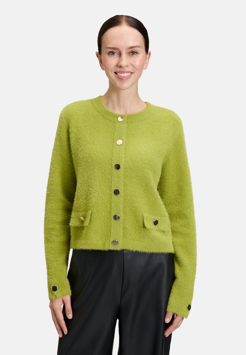 Une femme portant un cardigan boutonné pelucheux vert citron avec des poches et un pantalon noir, souriante, devant un fond blanc uni.