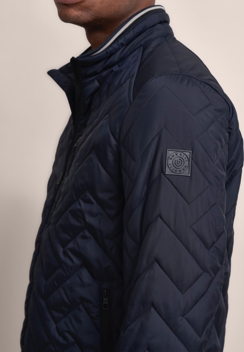 Navy gesteppte Jacke mit hohem Kragen, Zickzackmuster, seitlicher Reißverschlusstasche und einem Logopatch am Oberarm. Glatte Textur.