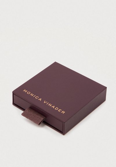 Hnedá štvorcová šperkovnica s textúrovanou stuhou na ťah a zlatým nápisom "MONICA VINADER" na prednej strane. Hladký povrch, kompaktný dizajn.