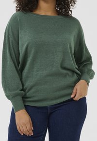 Groene, metallic gebreide sweater met een losse pasvorm, brede bootcollar en lange mouwen. Bevat ribgebreide boorden en een zoom, gecombineerd met donkere jeans.