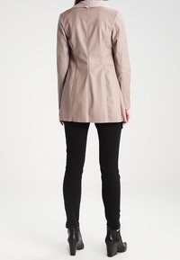 Abrigo de cuero beige claro con un diseño ajustado, cuello solapa y costuras decorativas, llevado sobre pantalones ajustados negros y botas de tobillo.