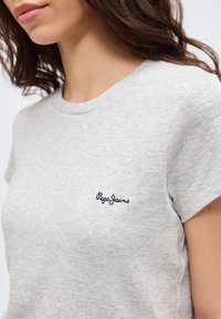 Gros plan d'une femme portant un t-shirt côtelé gris clair à manches courtes avec le logo "Pepe Jeans" brodé sur la poitrine.