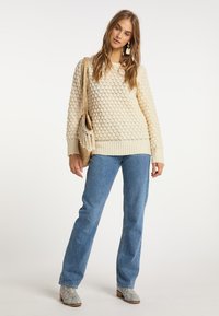 Krémszínű kötött pulóver texturált mintával, világoskék egyenes szárú farmernadrággal és mintás bokacsizmával párosítva. Fringes táskával kiemelve.