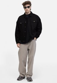 Chaqueta de denim negra con dos bolsillos frontales, llevada sobre una camiseta negra. Pantalones holgados gris claro y zapatillas grises completan el look.
