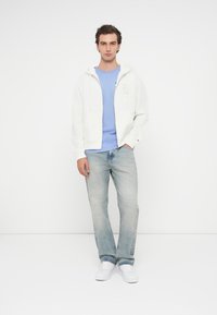 Λευκή φούτερ ζακέτα με τσέπες καγκουρό, σε συνδυασμό με ένα ανοιχτό γαλάζιο μπλουζάκι. Φορώντας φθαρμένο μπλε τζιν και λευκά αθλητικά παπούτσια.