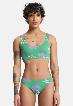 Person mit kurzen lockigen Haaren und mehreren Tattoos, die einen grünen Bikini mit Blumenmuster trägt und vor einem einfarbigen Hintergrund steht.