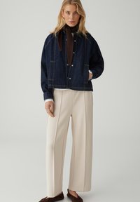 Woman wearing dark blue denim jacket, black scarf, cream wide-leg trousers, and brown shoes, standing against a plain light background.
