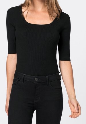 Kvinde iført sort ribbet top med albuelange ærmer og sorte bukser med høj talje, vist fra skuldre til hofter mod hvid baggrund.