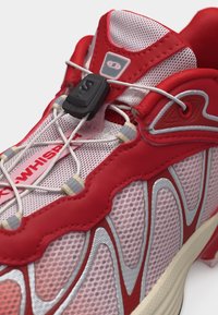 Κόκκινο και λευκό αθλητικό παπούτσι με σχεδιασμό από δίχτυ, ρυθμιζόμενα ελαστικά κορδόνια και μαύρο κλιπ στερέωσης στην περιοχή της γλώσσας.