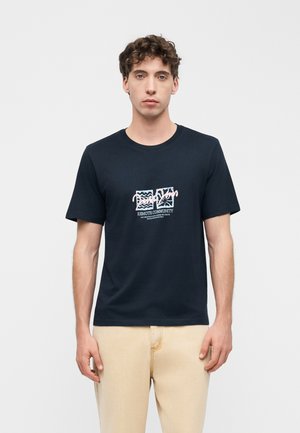 Jeune homme aux cheveux bouclés portant un t-shirt noir à motif graphique et un pantalon beige, debout devant un fond blanc uni.