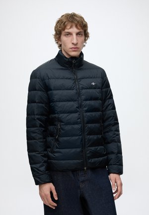 Junger Mann mit lockigem Haar, der eine schwarze gesteppte Jacke mit Reißverschluss und dunkle Jeans vor weißem Hintergrund trägt.