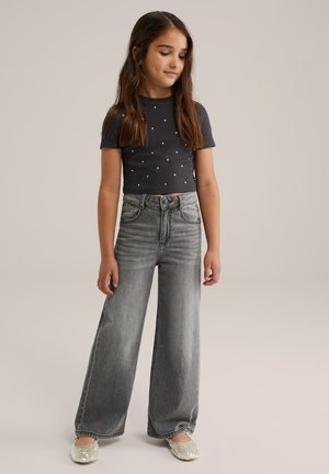 Graue Denimweite-Beine-Jeans kombiniert mit einem marineblauen Kurzarmtop mit weißen Punkten. Funkelnde Ballerinas runden das Outfit ab.