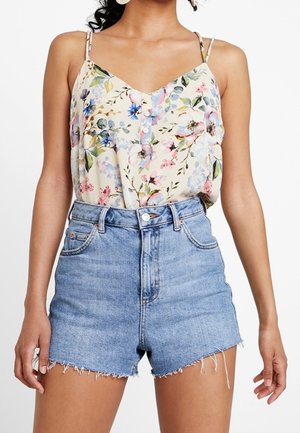 Žena nosí světle modré džínové šortky s vysokým pasem a roztřepenými lemy a krémový květinový top s tenkými ramínky a knoflíky.