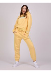 Gul hoodie og matchende sweatpants i blødt stof, med blå tekstdetaljer, ribbede manchetter og snørebånd i taljen. Båret med hvide sneakers.