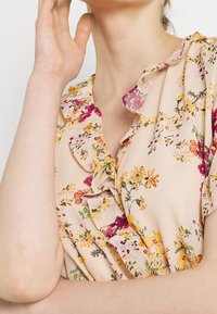 Kukkakuosiset pusero pehmeässä vaaleanpunaisessa kankaassa, jossa on röyhelökaulus ja elinvoimaisia keltaisia, vaaleanpunaisia ja vihreitä kukkakuoseja pinnalla.