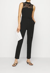 Mono negro con detalles de encaje en la parte superior, cintura ceñida y piernas ajustadas y afiladas. La modelo sostiene un bolso blanco y lleva sandalias negras.