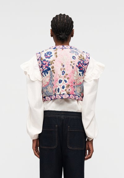 Personne aux cheveux tressés portant un chemisier blanc à manches volantées, un gilet sans manches à motif floral et un pantalon foncé avec des poches.
