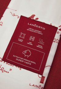 Czerwona naszywka z tkaniny z białym tekstem zawierająca instrukcje dotyczące pielęgnacji. Materiał: ekstra cienka wełna merino, super 140's. Cechy: elastyczny, odporny na zagniecenia, wodoodporny.