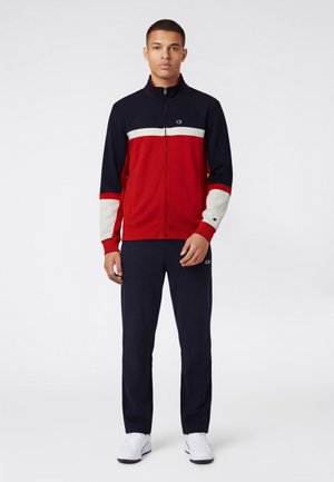 Hombre con chaqueta con cremallera azul marino, roja y blanca, pantalones azul marino a juego y zapatillas blancas, de pie contra un fondo liso.