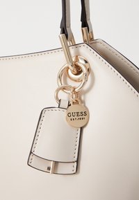 Borsa sintetica crema con hardware dorato, dotata di un'etichetta che recita "GUESS EST. 1981" e cuciture nere di contrasto lungo i bordi.