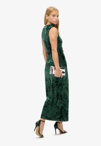 Robe de soirée en velours vert avec un design ajusté, sans manches et longueur longue. Présente une pochette argentée brillante et des chaussures à talons hauts noires.