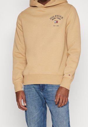 Hombre con sudadera beige con capucha con el texto "Hilfiger New York Est. 1985" y logo en el pecho, combinado con vaqueros azules.