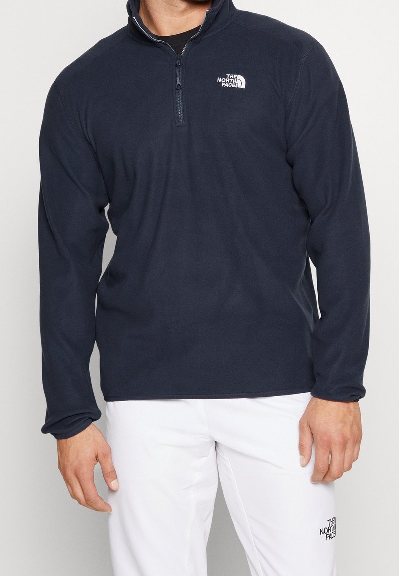 Homme portant une polaire bleu marine à demi-fermeture éclair et un pantalon blanc, tous deux avec le logo The North Face sur la poitrine et la cuisse.