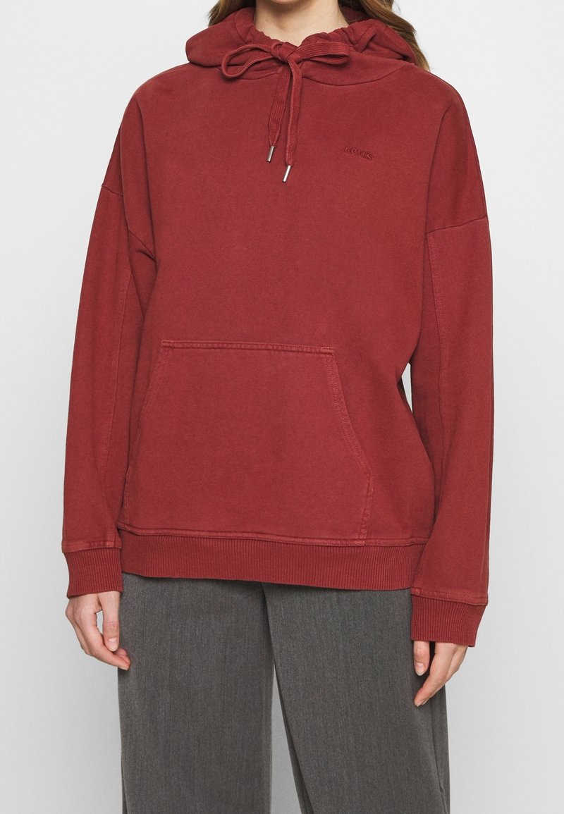 Persona vistiendo una sudadera roja holgada con cordones y bolsillo frontal, combinada con pantalones de pierna ancha grises contra un fondo liso.