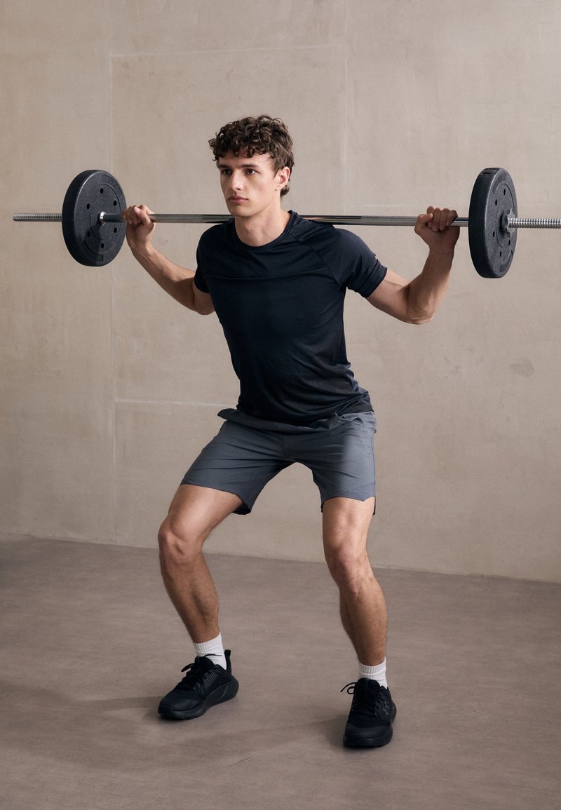 Camisa atlética negra y pantalones cortos grises usados por una persona en cuclillas con una barra. La barra tiene discos de peso negros. Entorno interior.