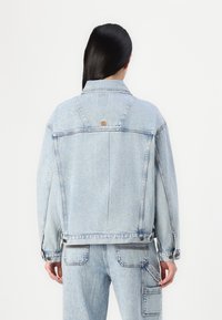 Chaqueta de mezclilla de lavado claro con un corte relajado, botones de metal y costuras dobles. Presenta un logo en la parte trasera y amplios bolsillos frontales.