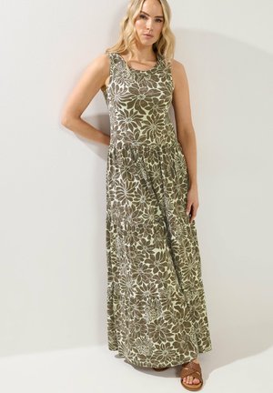 Blondynka w bezrękawnej oliwkowozielonej maxi sukience z dużym białym kwiatowym wzorem i brązowych plecionych sandałach, stojąca przy ścianie.