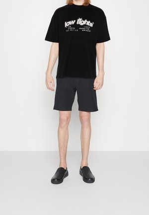 T-shirt en velours noir avec un texte graphique blanc, associé à un short sombre. Le modèle porte des baskets noires texturées, se tenant devant un fond uni.