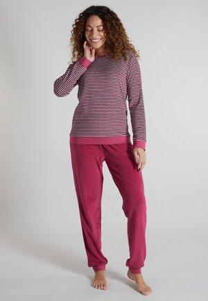 Woman with curly hair wearing a pink and white striped long sleeve top and solid pink pants, standing barefoot on a gray background.