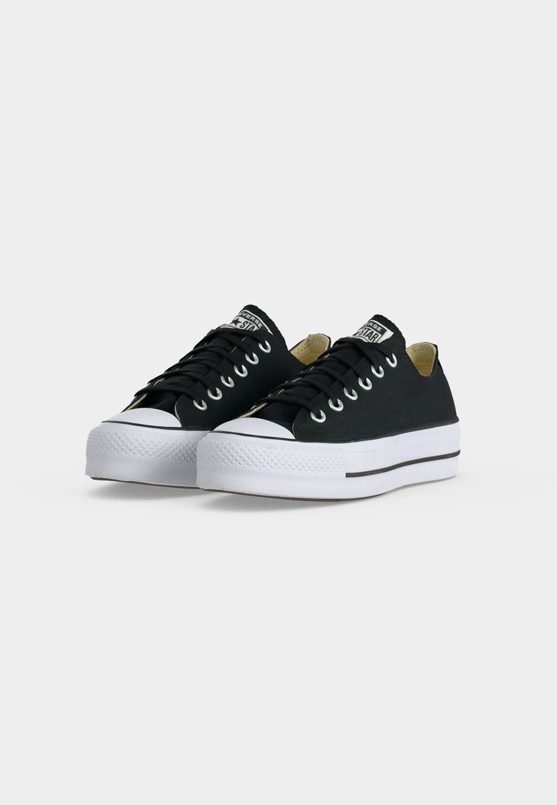 Coppia di sneakers basse nere in tela con suole e punte in gomma bianca, lacci neri e etichetta del marchio sulla linguetta su sfondo uniforme.