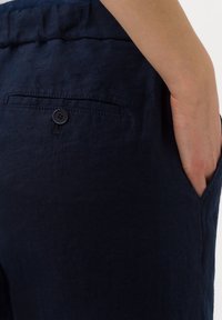 Pantalon en lin bleu marine avec une poche arrière équipée d'un bouton noir et une poche latérale, présentant une finition en tissu texturé.