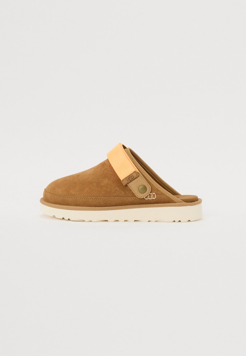 Tamancos de camurça castanhos, tipo slip-on, com uma faixa laranja claro e sola texturizada beje. Apresentam um bico redondo e um detalhe em botão lateral.
