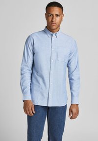 Camisa de botones de manga larga en azul claro, confeccionada en una tela texturizada, con un bolsillo en el pecho y cuello estándar, combinada con pantalones vaqueros oscuros.