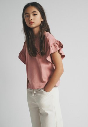 Hosszú, sötét hajú fiatal lány, aki poros rózsaszín fodros ujjú felsőt és törtfehér nadrágot visel, kezét zsebre téve, egyszínű háttér előtt.