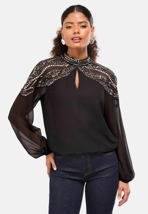 Žena u crnoj prozirnoj bluzi s dugim rukavima, ukrašenoj perlicama na visokom ovratniku i detaljima na ramenima, u kombinaciji s tamnoplavim trapericama.